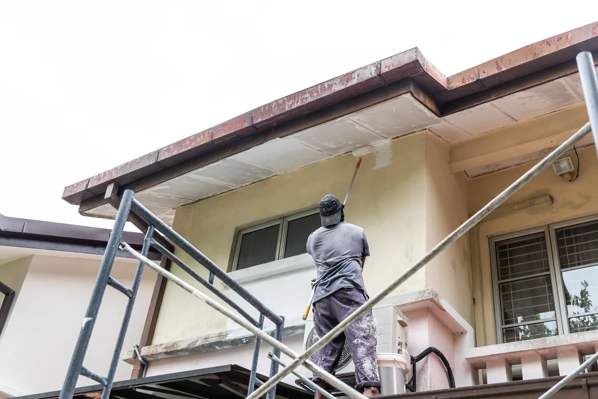 Painter applying fresh paint to a two-story stucco exterior from scaffolding