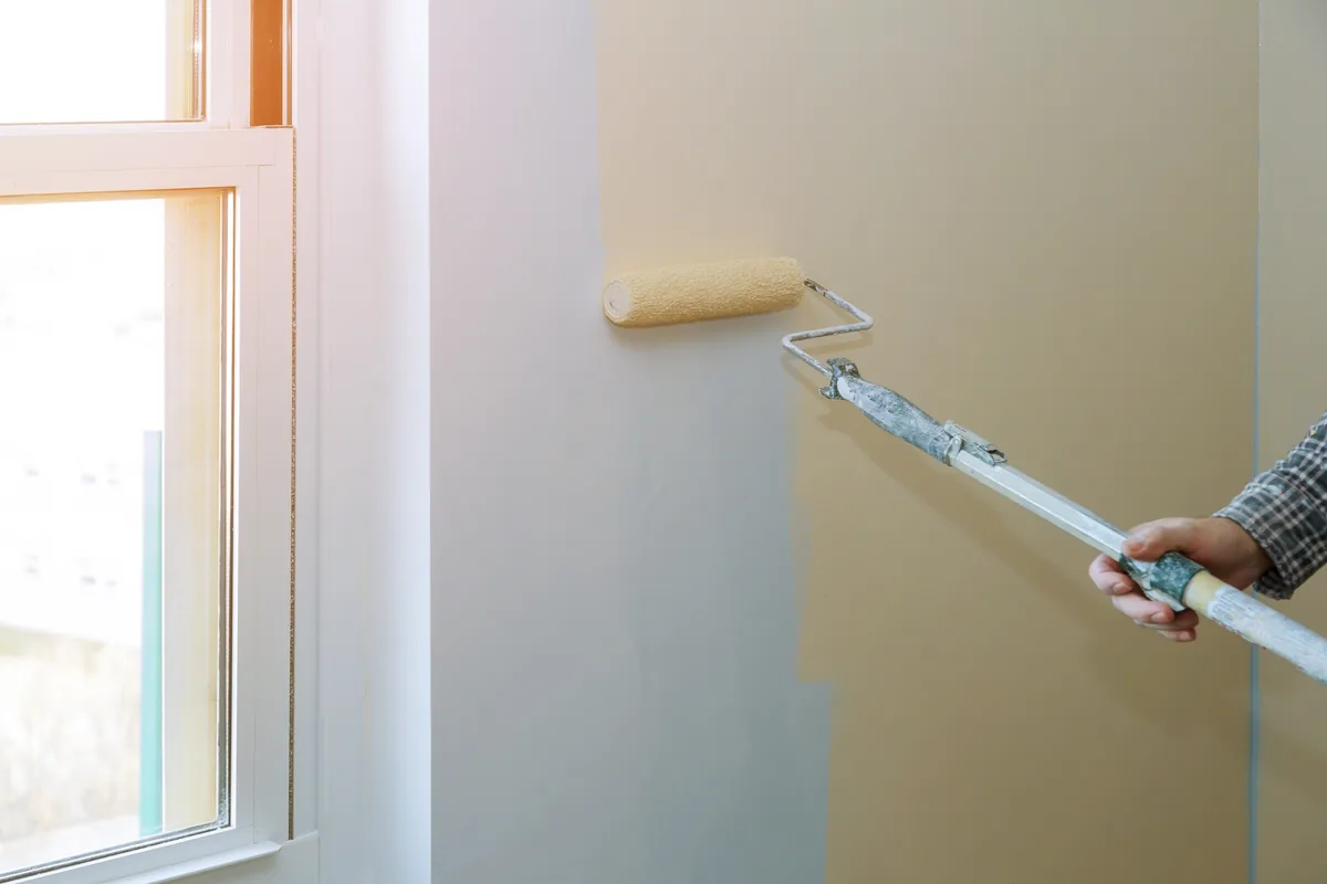 Roller applying paint to an interior wall beside a window